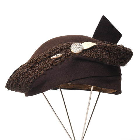 antique 1920s brown felted hat with faux astrakhan trim with wider flared sides, angled flared up back piece and cream leaf and circle rhinestone celluloid center front hat pin shown on four metal thin leg hat stand against white background
