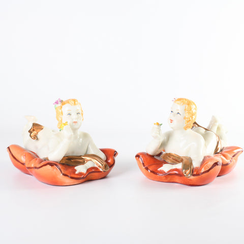 2 porcelain vintage piano babies lying on stomachs on porcelain orange pillow holding flower in right hand looking up shown angled toward each other on white background