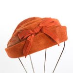 true vintage orange felted fur tilt hat with felt folded to create 3 tiers finished with front center orange bow and feather behind it that runs behind the turned up brim shown on thin silver metal 4 leg hat stand all on white background