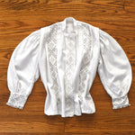 genuine antique early 1900s white hardanger embroidery traditional bunad blouse with wide balloon sleeves shown lying flat on warm brown hardwood floor