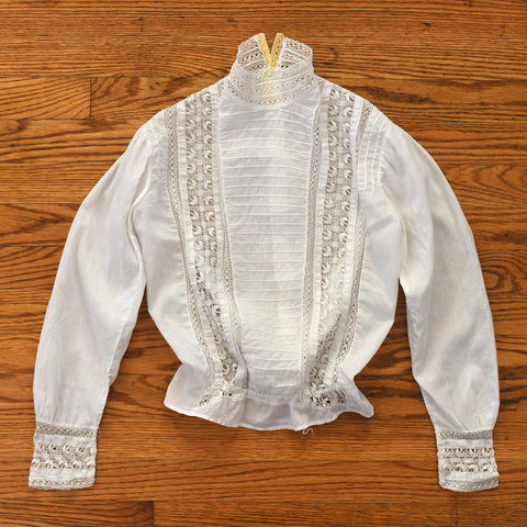 early 1900s white cotton pigeon chest blouse with hardanger embroidery butterflies running vertically, pin tucks running down the center of the blouse horizontally and trimmed with lace shown lying on warm brown hardwood floor