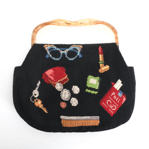 true vintage mid 1950s to mid 1960s black needlepoint handbag with novelty print theme of purse contents including blue sparkly cat eye glasses, spilled coins coin purse, keys, hair comb, lipstick, match book and Pall Mall cigarettes with clear and brown flip top acrylic handle shown on white background