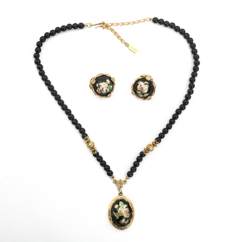 true vintage 1980s black beaded necklace with gold tone latch hook closure and black floral enamel oblong center pendant with circular matching enamel earrings sitting in center of necklace shown on white background