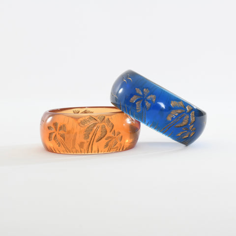 true vintage translucent orange carved gold rubbed palm tree bangle lying flat with translucent blue carved gold rubbed palm tree bangle angled sideways against orange bangle right side of image all against white background