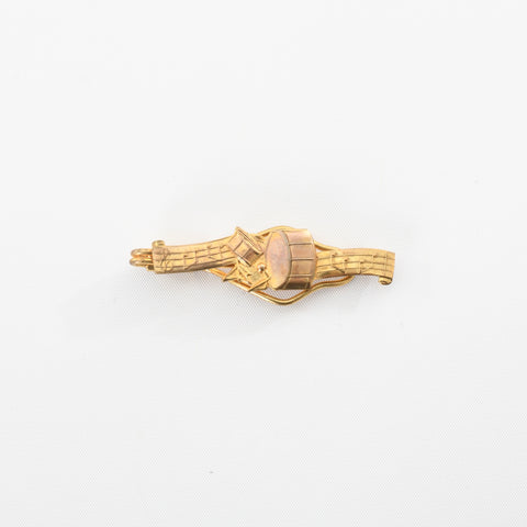 true vintage 1950s gold toned tie bar with musical staff seperated by a snare drum and kick drum shown lying flat on white background