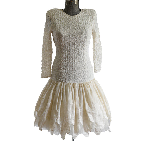 cream lace dropped waist long sleeve dress with full ivory petal hem full midi skirt with tulle showing at bottom all on dress form against white background