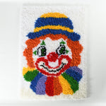 vintage 1970s latch hook clown rug with rainbow collar, orange hair, smile, blue and yellow hat on a white background