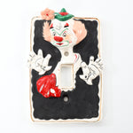 vintage 1950s ceramic clown wall light switch plate with peach hair, green hat and peach flower appearing like he pops out of dark with his hands up shown on white background